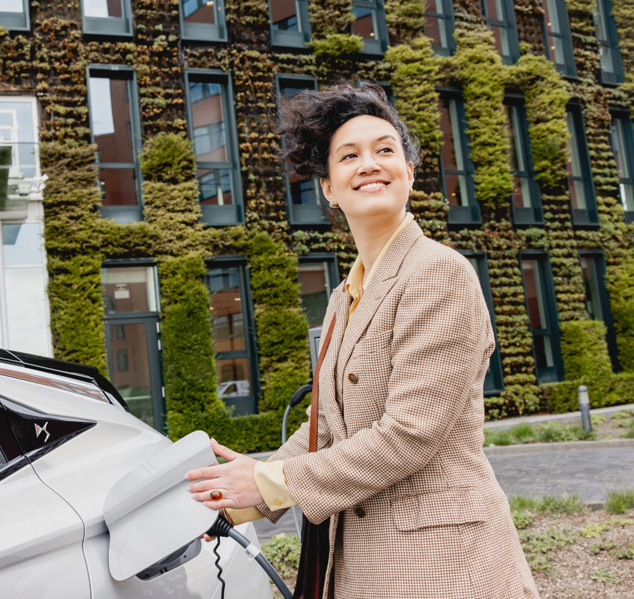 Woman in a business suit stands next to a white electric car parked in front of a modern building with a green facade.