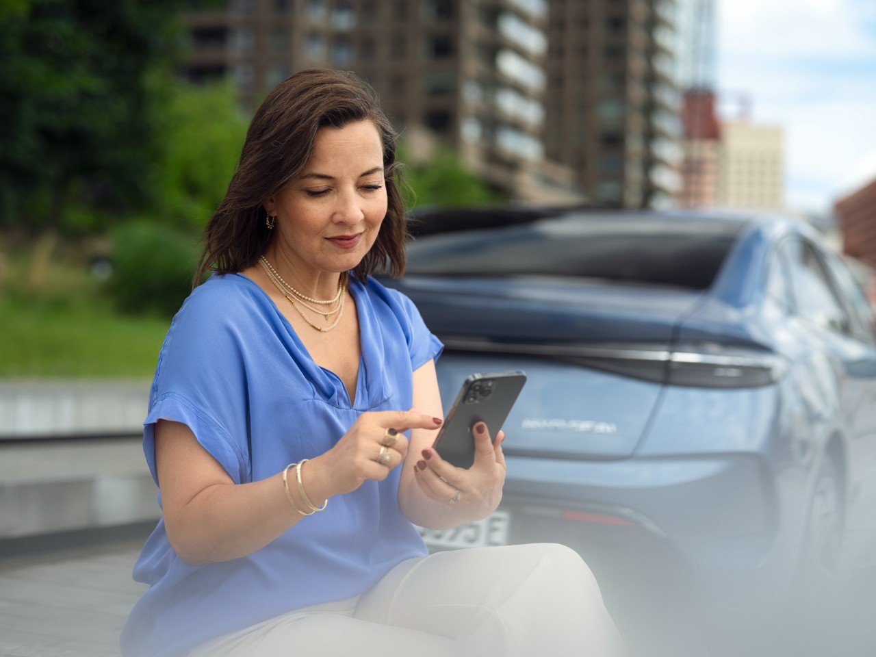 A woman uses her phone while the rear of a blue-grey car is visible in the background.
