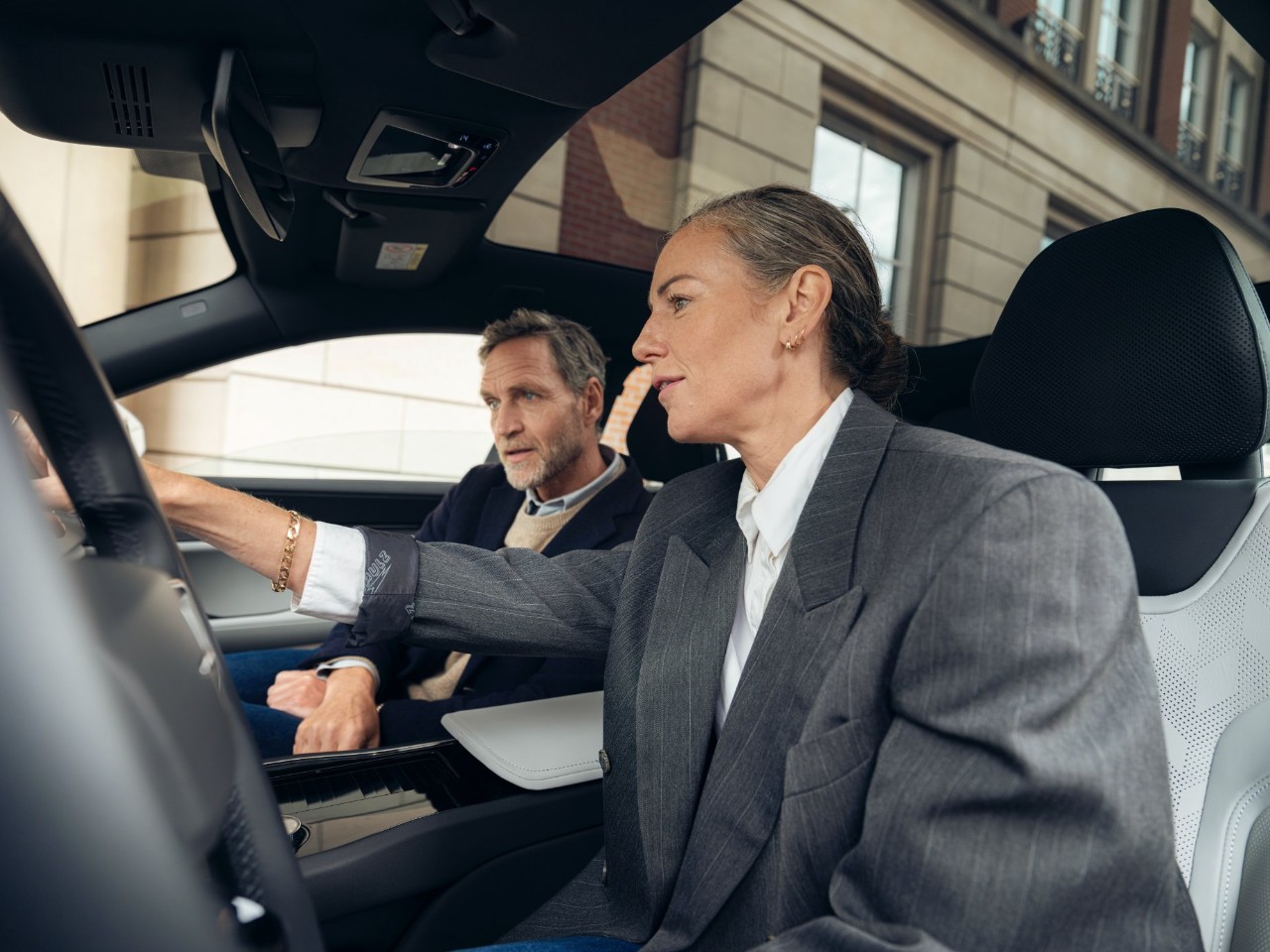 Two people in blazers sit in a car and look at the central display.