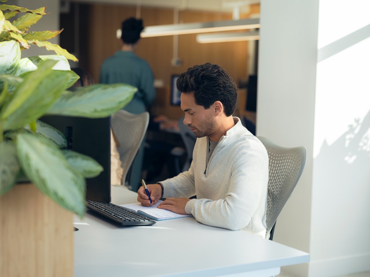 Man in a white sweater sits at a computer and takes notes in a notebook.