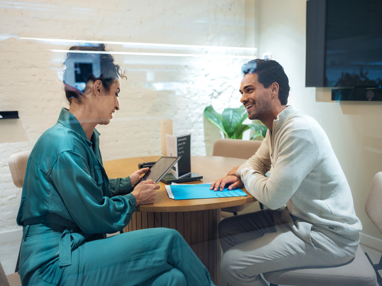 Two people sitting at a round wooden table in an office, discussing documents and a tablet. 