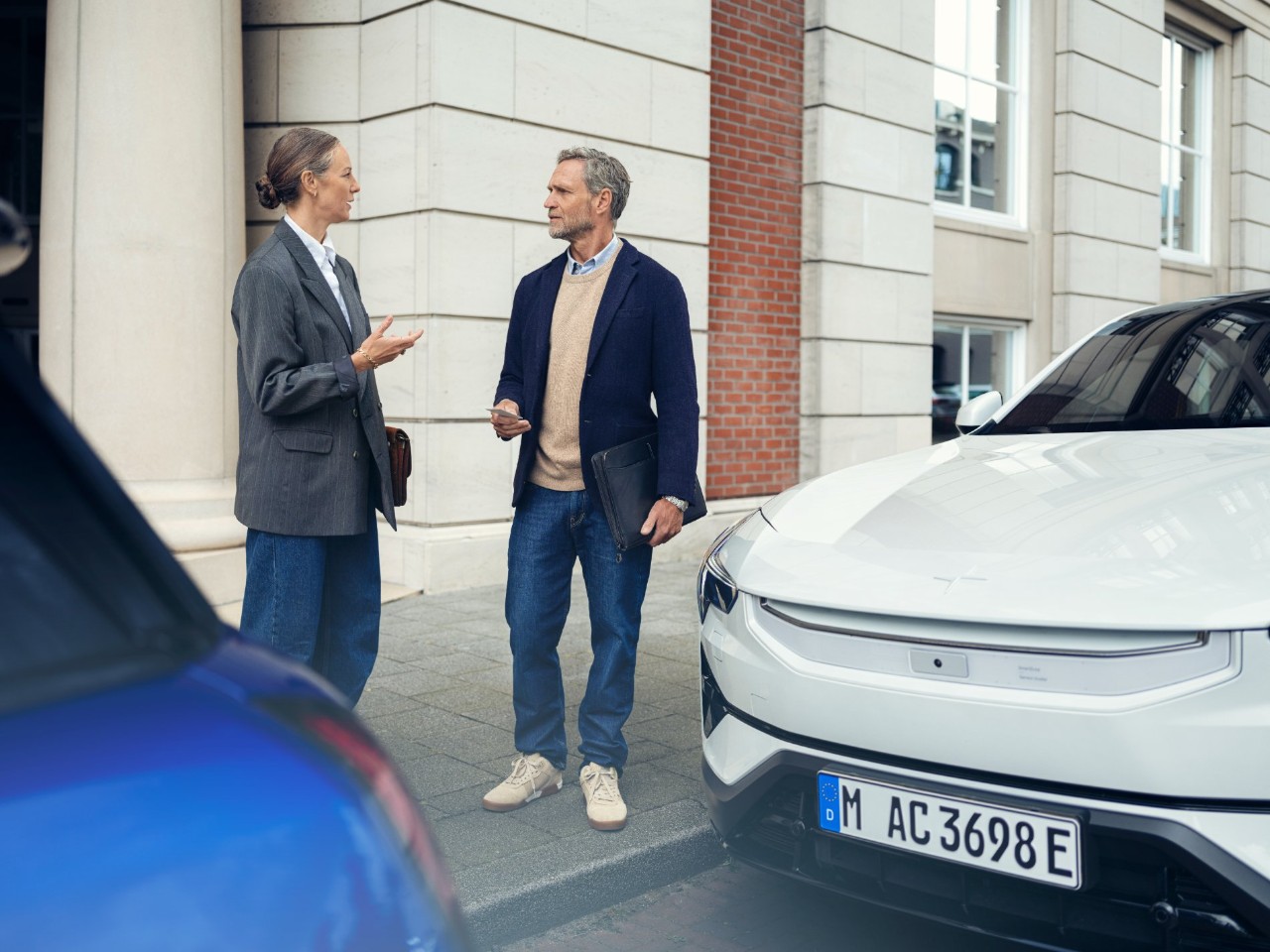 A woman and a man have a conversation on the sidewalk in front of an elegant building, next to two parked cars.