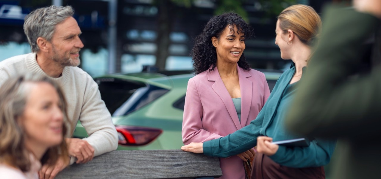 Three women discuss in front of a green car, holding a tablet. 