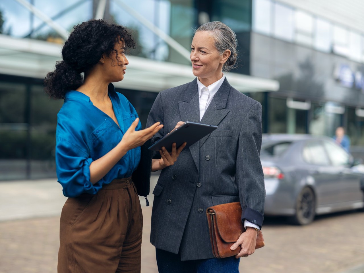 Two women have a conversation in front of a building with large glass windows.