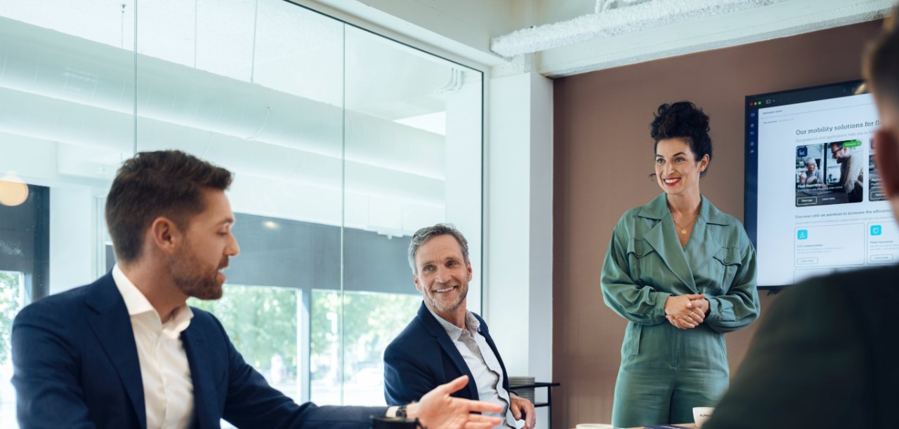Three people are having a discussion at a wooden table in a modern conference room with a large screen.
