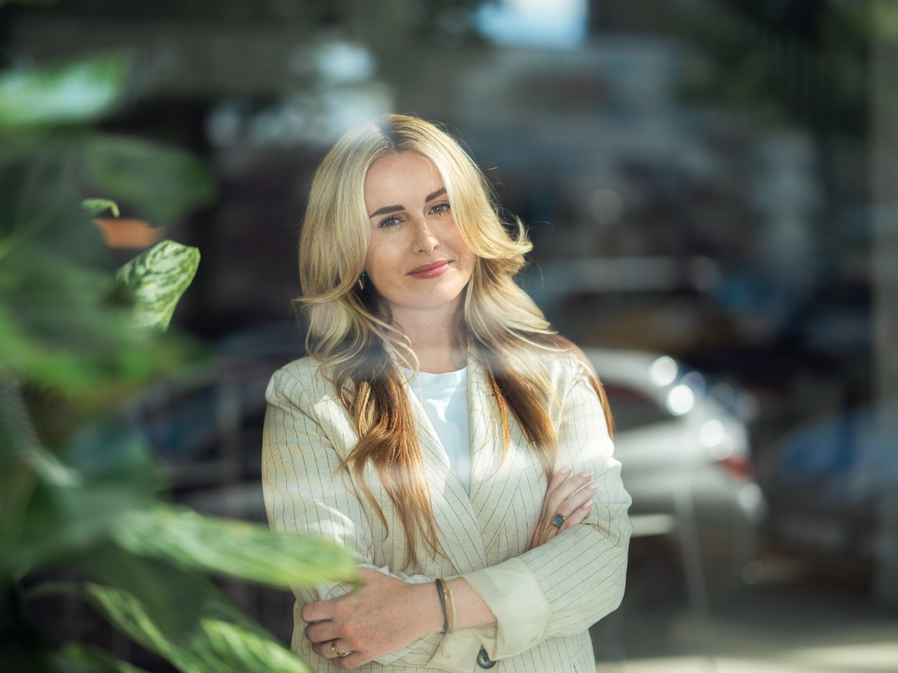 Woman with arms crossed smiling looking directly out with green plant blurred out to the left