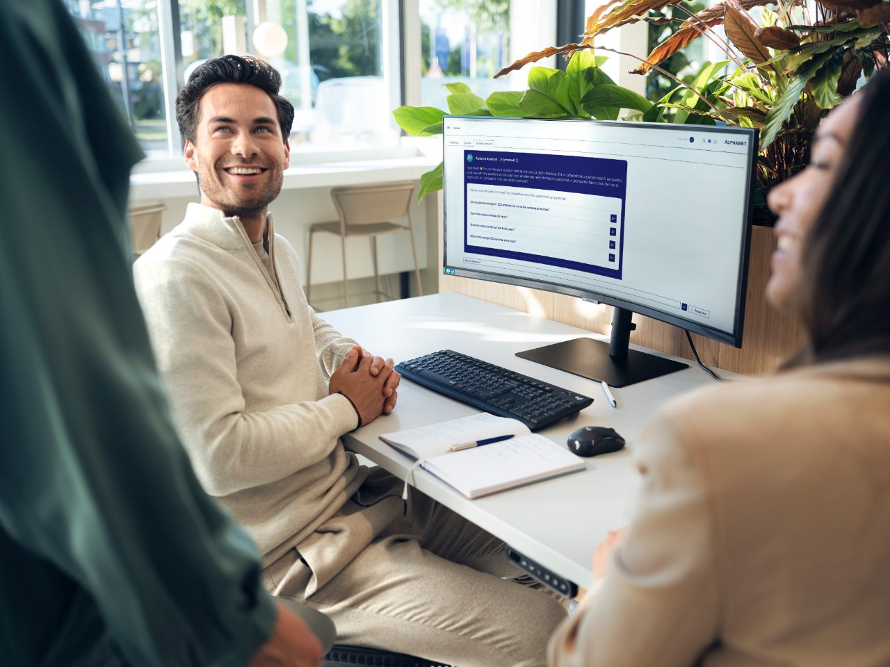 Person sitting at a desk in a modern office, working on a computer with the Alphabet website open, surrounded by green plants and large windows.
