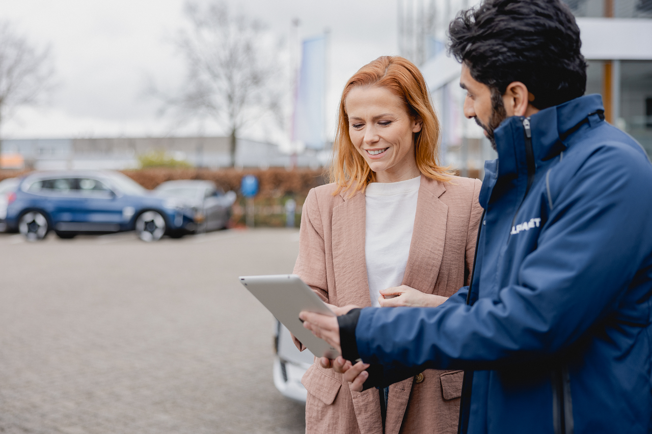 Two people standing outdoors next to a car, discussing information displayed on a tablet.
